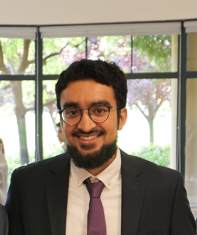 Headshot of Afras smiling, wearing a black suit and black rimmed, clear lens glasses